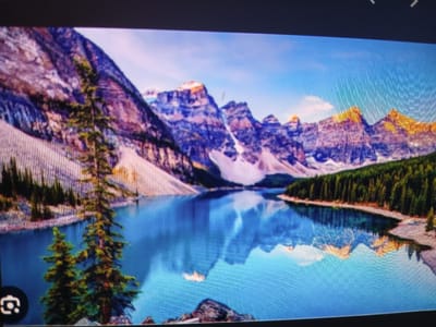 The image is a scenic landscape photograph of a tranquil lake reflecting majestic mountains and a dense forest. The mountains, tinged with purple and gold hues, rise sharply against a clear blue sky. Lush green pine trees line the shore, their reflections mirrored in the calm, blue waters of the lake. In the foreground, a few rocks and evergreen trees add depth to the composition. The overall impression is one of serene natural beauty. This image is not located in Siheung-si, South Korea.