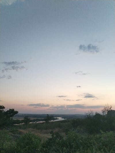 A wide-angle, low-angle landscape photograph captures a serene twilight view over the Sula River in Bilousivka, Ukraine. The sky transitions from a light blue at the top to a soft pinkish hue near the horizon, with scattered wispy clouds. The foreground is dominated by dense, dark green foliage and dry grasses, suggesting a natural, somewhat overgrown terrain. Beyond this, the Sula River winds its way through a broad valley, reflecting the muted colors of the sky. Distant fields and trees form a layered landscape that recedes towards the horizon. The overall impression is one of natural beauty and tranquility at dusk.