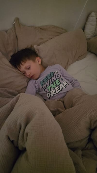 A child, wearing a lavender long-sleeved shirt with the text "KEEP DREAMING" in colorful lettering, is sleeping on a bed. The bed is covered with a beige blanket, and there are pillows in the background. The location of the image is not provided.