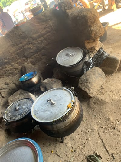 The image depicts an outdoor scene in Dull A, Nigeria, likely a cooking area. Several large, dark metal pots with silver lids are arranged on the ground, some resting on rocks or a rustic stove-like structure made of earth. One pot to the right is partially open, revealing what appears to be food inside. Another smaller, dark pot with a blue interior is nestled between rocks. The ground is a mixture of sand and dirt. In the background, the scene suggests a marketplace or a communal area, with hints of wooden structures, fabric, and possibly people blurred by motion or distance. The lighting suggests it could be daytime, with sunlight casting shadows. The overall impression is one of everyday life and food preparation in a rural or semi-rural setting. No text is visible in the image.