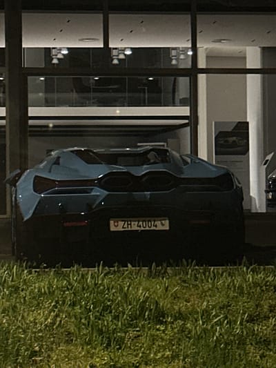 A blue car with license plate "ZH-4004" is parked next to a grassy area. A building with exterior wall-mounted lights and windows is visible behind the car. The license plate suggests the location is in the canton of Zurich, Switzerland. Kloten is a city in that canton.