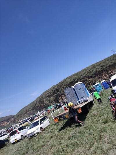 A wide-angle, tilted photograph captures an outdoor gathering in Olkaria ward, Kenya, under a clear blue sky. The scene is dominated by a large, grassy field that slopes gently upward from the foreground toward a steep, heavily vegetated hillside in the background.
In the immediate mid-ground, a white flatbed truck is parked facing away from the viewer, carrying two grey portable toilets with white doors. License plates are partially visible on the truck's rear, along with a red '80' sticker. To the right of the truck, a person wearing a dark shirt, dark pants with red stripes, and a yellow protective helmet or hat walks left across the grass, carrying a yellow container in their right hand. Their shadow is visible on the grass to their left.
Numerous other vehicles, including several white cars (one with license plate KCH 829K) and motorcycles, are parked on the grassy field, extending into the mid-ground and background. A large crowd of people is gathered further up the slope, many individuals wearing green apparel. More portable toilets, some blue and white, are visible among the crowd near the base of the hillside. High-voltage power lines and a pylon structure are discernible on the distant, tree-covered ridge to the right. The overall environment suggests a sunny day with distinct shadows cast by subjects and objects.