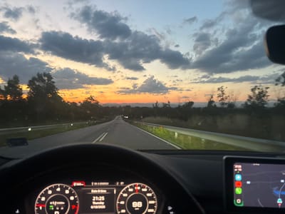 The image depicts a view from the driver's seat of a car, driving on a road at dusk. The sky is filled with dramatic clouds tinged with orange and yellow from the setting sun. The road ahead is visible, with a guardrail on the right side and trees lining the landscape. The car's dashboard is in the foreground, showing the speedometer at 98 km/h and the date and time as 21:25, Thursday, 6/18/2020. A navigation screen is also visible on the right, displaying a map. The location is identified as being in the vicinity of Aleko Konstantinovo, Bulgaria.