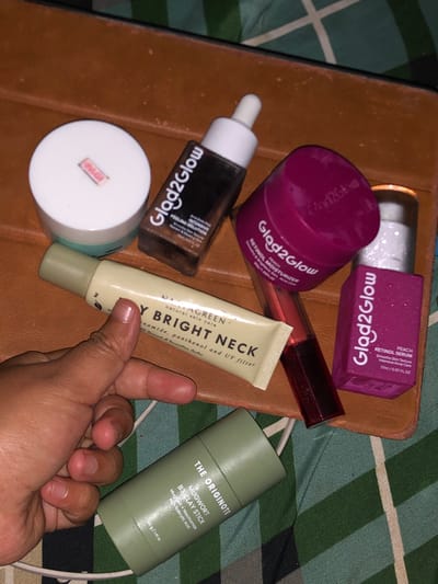 An indoor, close-up, eye-level image captured in Tlogo, Indonesia, displays a collection of cosmetic and skincare products arranged on a rectangular brown surface, consistent with a leather tray or case. A human hand, exhibiting medium-dark skin pigmentation, occupies the lower-left quadrant, holding a light green tube labeled "NATURAGREEN DAY BRIGHT NECK" with a gold cap. Other visible items include: a dark brown "Glad2Glow AHA BHA PHA PEELING SOLUTION" dropper bottle with a white cap, a magenta "Glad2Glow RETINOL MOISTURIZER" jar with a matching lid, a magenta rectangular "Glad2Glow PEACH RETINOL SERUM" bottle with a white cap, an olive green "THE ORIGINOTE MUGWORT B3 CLAY STICK," a white cylindrical jar with a light blue base and a small pink label (partially obscured, potentially "PAGI"), and a clear, cylindrical cosmetic item with a dark cap containing a reddish-brown liquid. The immediate background reveals a green and white checkered or plaid fabric pattern, indicative of a bed linen or similar textile surface.