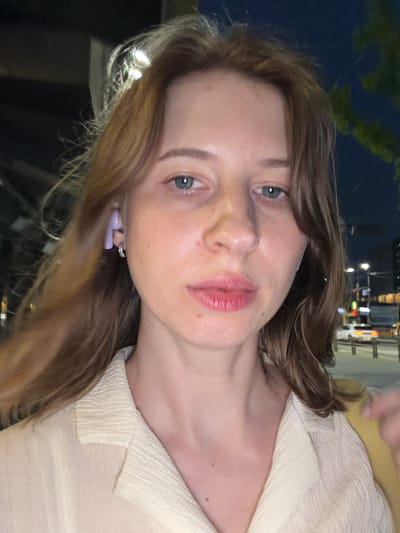 A young Caucasian woman with shoulder-length, light brown hair is captured in a close-up, selfie-style shot at night in Uijeongbu-si, South Korea. She is wearing a light-colored, textured collared shirt and has a purple earbud in her left ear. Her face is the central focus, with a neutral expression. The background suggests an urban environment at night, with blurred lights from street lamps and buildings visible. A few vehicles and street barriers are faintly discernible in the distance, indicating a city street or thoroughfare. The lighting is dim, consistent with nighttime conditions.