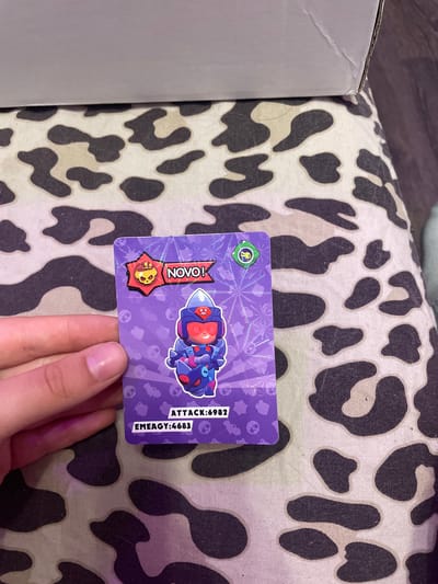 A Brawl Stars collectible card is held by a person's hand against a backdrop of a black and white cow-print fabric. The card is predominantly purple with holographic accents. At the top left of the card is a star-shaped icon with a skull and a golden outline, next to the word "NOVO!" in a banner. A stylized character resembling an armored figure with a red visor is depicted in the center of the card. Below the character, text reads "ATTACK: 6982" and "EMEAGY: 4683". The environment is a close-up indoor shot, with no visible indications of the city of Vaganovo Massiv, Russia.