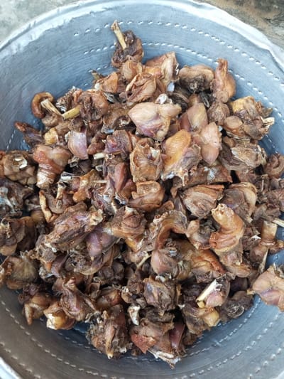 A close-up, high-angle shot shows a metal bowl filled with cooked chicken pieces. The chicken appears fried or roasted and has a brown color. Some pieces show visible bones. The bowl has a gray, metallic appearance with concentric ring patterns and small perforations. The image has a slightly grainy texture. This image was taken in Isunba, Nigeria.