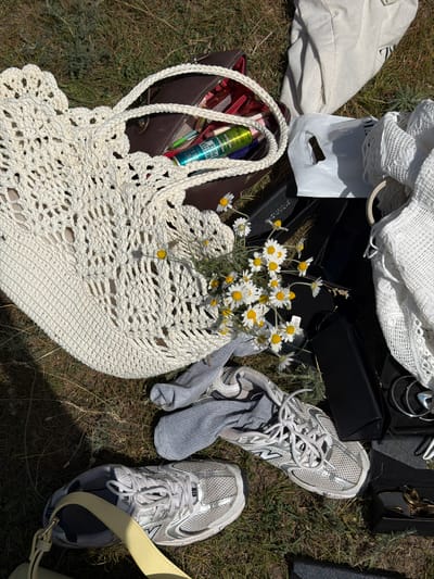 The image captures a collection of personal belongings scattered across dry, grassy ground in bright daylight, with a backdrop consistent with an outdoor setting in Баш-Кара-Суу, Kyrgyzstan.
**Subjects:** A central element is a large, open-weave, cream-colored crocheted bag, from which a bouquet of white and yellow daisies spills out. Adjacent to it, a dark brown leather handbag is open, revealing items inside, including a green cosmetic tube. A pair of white and silver New Balance sneakers with gray socks tucked into one rests on the grass. Other objects include a crumpled white canvas tote bag, a white plastic shopping bag, a white lace garment or fabric, and several black cases or wallets. Various accessories, such as silver hoop earrings, other jewelry, and keys, are also visible. A light yellow strap from another bag extends into the frame.
**Activity or event taking place:** The scattered arrangement of items suggests a casual moment of unpacking, repacking, or taking a break during an outdoor activity, such as a picnic, a hike, or a relaxed outing. The freshly picked daisies imply a connection to nature.
**Notable details:** The time of day appears to be sunny daytime, with clear, bright light casting shadows. The weather is clement and sunny. The ground is dry grass with some sparse wild plants. The overall atmosphere is relaxed and informal.
**Visible text:** On the green cosmetic tube inside the brown bag, the words "MASTER PIECE" are legible, along with smaller product details. The "N" logo for New Balance is clearly visible on the side of the sneakers. Text on the white plastic bag is present but not decipherable.