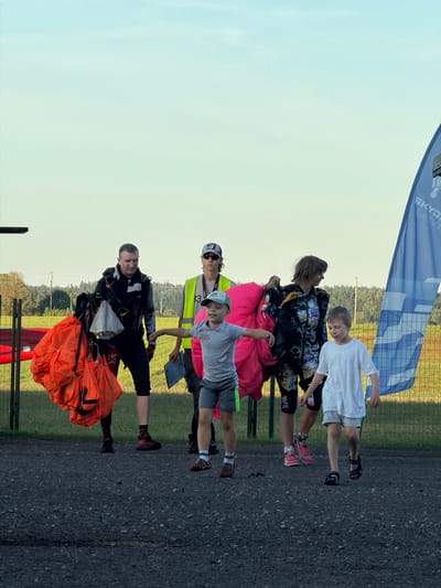 A group of people, including adults and children, are gathered outdoors in what appears to be an airfield or similar open space. A man on the left is wearing a dark jumpsuit and carrying a large orange parachute. Next to him, a person in a neon yellow vest and a cap stands with a clipboard. To the right of them, a woman is dressed in a colorful jumpsuit and is holding a large pink parachute. Two young boys are also present; one in a gray t-shirt and shorts is looking to the left, while another in a white t-shirt and gray shorts is walking forward. A blue banner with white lettering, partially visible and bearing the word "SKYDAY", is positioned on the right side of the frame. The background consists of green fields and trees under a clear sky. The scene suggests a skydiving or parachuting activity. The mention of Limbaži, Latvia, implies this location is where the event is taking place.