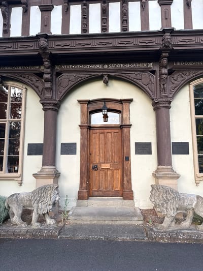 The image shows the entrance of a historic building in Leominster, United Kingdom. The building features dark brown timber framing against a light-colored facade, with decorative carvings above the arched doorways. Two stone lion statues stand guard on either side of a central wooden door, which has a lantern hanging above it. Plaques are visible on the walls next to the entrance, one reading "QUEEN ELIZABETH II" and another mentioning "HRH PRINCESS MARGARET." The ground in front of the building is paved, with a section of asphalt road in the foreground. Green foliage is present around the lion statues.