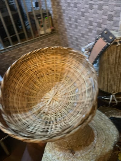A close-up shot showcases several woven baskets. A round basket takes center stage with its intricate, spiraling weave pattern. Additional baskets, including a rectangular one with a leather strap and a straw hat, are partially visible in the background. The scene includes glimpses of a gray, patterned surface and a metal rack with horizontal bars. The overall impression is of a collection of handcrafted, natural fiber objects.