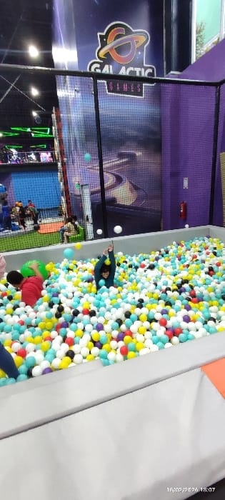 A child in a teal shirt lies in a ball pit filled with white, yellow, blue, red, and black balls. The pit is surrounded by gray walls and an orange section. A play area with nets, arcade-style games, and a Galactic Games logo is visible in the background. Other individuals are seen in the distance. The image is rotated, and a timestamp is in the upper right corner.