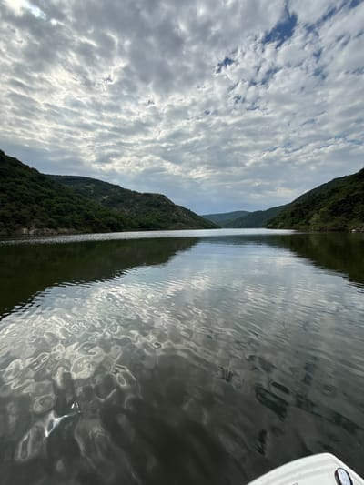 The image shows a body of water, possibly a lake or river, with a forested mountain range on one side and a cloudy sky reflected in the water. A small part of a white boat or object is visible in the upper right corner. The location is likely Senoklas, Bulgaria, based on the context. No people are visible. The primary subject is the landscape, focusing on the water, mountains, and sky.