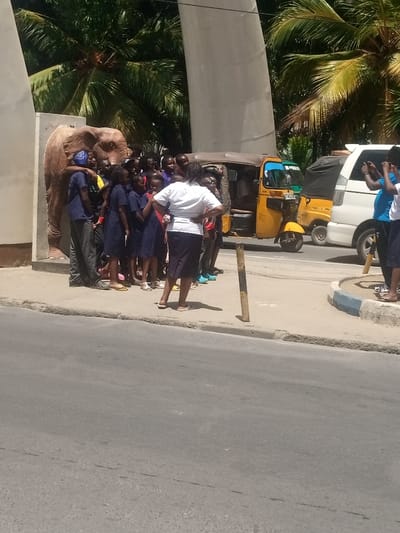 An outdoor daytime scene in Ganjoni/Shimanzi ward, Kenya, depicts a group of approximately 15 to 20 individuals, predominantly children wearing dark blue school uniforms, gathered on a paved concrete sidewalk. They are positioned around a prominent architectural monument consisting of large, pale structures resembling elephant tusks, a known landmark at Moi Avenue. A brown sculpted elephant head is visible at the base of the left tusk. An adult female, dressed in a white short-sleeved top and dark skirt, stands centrally with her hands on her hips, facing away from the camera, seemingly supervising the group. An adult male wearing a blue shirt and head covering stands to the far left, near the elephant sculpture. Several individuals are observed interacting with or posing near the sculpture. On the far right, two individuals are visible, one holding a mobile phone in a photographic posture, standing near a blue and white painted road curb. On the asphalt road behind the group, a yellow three-wheeled auto-rickshaw (tuk-tuk) and a white van are parked or stationary. The background features dense green palm tree foliage under bright, clear daylight conditions.