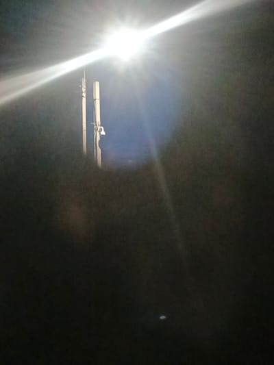 The image depicts a night scene illuminated by a bright overhead light source, likely a streetlamp, positioned in the upper portion of the frame. The light casts a strong glare and rays, creating a hazy effect. In the mid-ground, two vertical poles are visible, one to the left and one to the right, appearing to be part of a lighting or utility structure. A small, white object, possibly a camera or a fixture, is attached to the right pole. The background is dark and indistinct, suggesting an outdoor environment at night, possibly a street or open area within Воскресенское, Russia. The overall impression is one of low visibility and artificial illumination.