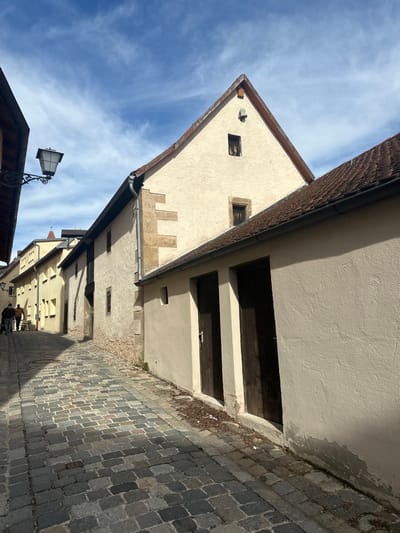 The image displays a narrow, cobblestone street in Weißenburg in Bayern, Germany, receding into the distance under a bright blue sky with scattered cirrus clouds. On the left, an ornate black street lamp is affixed to a building facade. Further along this side of the street, a line of light-colored, multi-story buildings features various windows and a prominent downspout. Two individuals are observed in the background on the left: one appears to be pushing an object on wheels, while the other walks nearby. The street itself is paved with multi-toned gray and brown cobblestones, with a distinct shadow line extending from the buildings on the right. The right side of the scene is dominated by a long, light-beige wall containing two dark wooden doors at ground level, each with a silver handle. Above and behind this lower wall, a larger, cream-colored building with a steep, reddish-brown tiled gabled roof stands prominently, displaying two small, dark-framed windows on its upper gable facade.