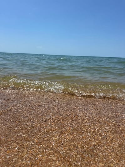 A low-angle, eye-level shot captures a pristine beach with gentle waves lapping at the shore under a clear blue sky. The foreground is dominated by a sandy beach composed of small, colorful pebbles and shell fragments. A thin, foamy wave, shimmering with sunlight, breaks over the pebbles, creating a dynamic line between the wet and dry sand. The ocean stretches out in the mid-ground, its surface rippling with a subtle texture. The horizon line is sharp, separating the calm sea from the vast, cloudless blue sky that fills the upper portion of the image. The scene appears to be a tranquil day at the beach in Blagoveschenskaya, Russia.