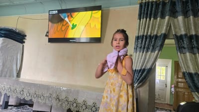 A young girl stands in a room, looking towards the camera with a surprised or curious expression. She is wearing a yellow dress with a floral pattern and has a light pink cloth wrapped around her neck, which she is adjusting with her hands. To her left, a large television screen is mounted on the wall, displaying a colorful animated image with the "BINGE SOCIETY" logo visible in the top left corner.
The room appears to be a domestic setting, possibly a living room or dining area. A long table covered with a clear plastic tablecloth is in the foreground. The table is adorned with an ornate lace trim along its edges. To the far left of the image, a stack of folded textiles, some appearing to be blue and white, are placed on a surface.
Behind the girl, a doorway is framed by decorative curtains with vertical stripes of dark blue, grey, and silver. Through the doorway, another room is visible with bright green walls and wooden doors. A window with a white frame and glass panes can be seen in the background, suggesting it is daytime. The overall lighting suggests it is likely daytime.
The style of the curtains and the general decor hint at a traditional or modestly furnished home. The presence of the television and the girl's attire suggest a contemporary scene. The image captures a candid moment, with the girl's expression and action of adjusting the cloth giving a sense of spontaneity. The location context of Yakkabog, Uzbekistan, implies a Central Asian setting.
On the television screen, the text "@Universal Pictures" is faintly visible at the bottom right.