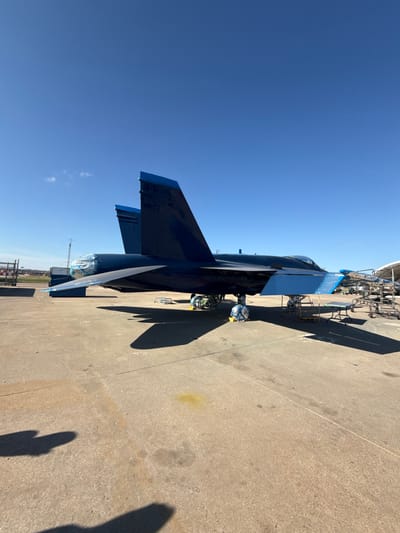 A dark blue fighter jet sits on concrete under a bright blue sky. The aircraft has two vertical stabilizers and two horizontal stabilizers. Scaffolding and wheeled carts are visible behind the jet. The nose and tail are covered with a protective wrap. Shadows from the aircraft and objects fall on the concrete surface.