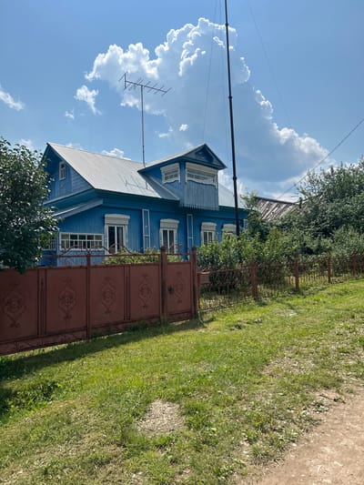 The image depicts a bright, sunny day in Новомуллакаево, Russia, with a clear blue sky dotted with fluffy white clouds. A two-story, blue wooden house with a corrugated metal roof stands prominently. The house features white-trimmed windows, some with decorative shutters. A dark red, ornate metal fence runs across the foreground, separating the property from a grassy area. Behind the house, lush green trees and foliage are visible. An outdoor television antenna is mounted on a pole on the roof of the house. A tall, dark pole with wires extends upwards in the right side of the frame. The overall scene suggests a residential setting in a rural or semi-rural area of Russia.