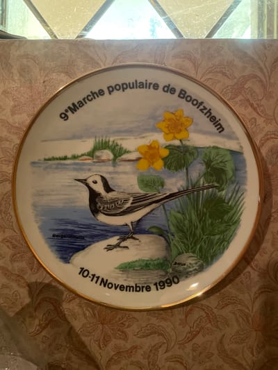 This is a round decorative plate featuring a painted image of a white wagtail bird perched on a branch over water, with yellow flowers and green foliage in the background. The plate has text inscribed around its circumference. On the left side, curving upwards, is the text "9. Marche populaire de Boofzheim". On the right side, curving downwards, is the text "10-11 Novembre 1990". A small inscription near the bird reads "Bargeronhatta". The scene depicts a natural setting, possibly a riverside or marsh area, with a bright, clear blue sky and water. The lighting suggests daytime. The plate appears to be a commemorative item, likely marking the 9th popular market event in Boofzheim, France, which took place on November 10th and 11th, 1990. The plate itself is bordered by a gold rim. The background of the image shows a patterned wallpaper.