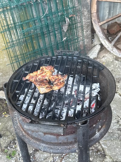 A high-angle shot depicts a piece of meat grilling on a round, charcoal grill. The grill is positioned outdoors on a stone surface. Partially burned charcoal is scattered on the grill beneath the meat. To the left of the grill is a section of green wire mesh fencing, and in the upper left corner is a portion of a wooden wagon wheel. The location is likely Boltata, Bulgaria, based on image context.
