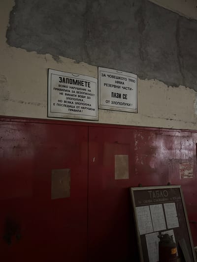 This media file displays an indoor scene, likely within an old industrial or utility building in Bratsigovo, Bulgaria. The image is dominated by a textured, peeling concrete or plaster wall on the left, and a distressed, dark red metal door or cabinet on the right. The overall impression is one of age and wear.
No human subjects are present, nor is any direct activity depicted. The scene is static, focusing on informational signage.
Notable details include two prominent safety signs attached to the wall, and a bulletin board mounted on the red surface. The signs are in Bulgarian.
The upper right sign reads:
"ЗА ЧОВЕШКОТО ТЯЛО НЯМА РЕЗЕРВНИ ЧАСТИ- ПАЗИ СЕ ОТ ЗЛОПОЛУКИ!"
(FOR THE HUMAN BODY THERE ARE NO SPARE PARTS – PROTECT YOURSELF FROM ACCIDENTS!)
The lower left sign states:
"ЗАПОМНЕТЕ ВСЯКО НАРУШЕНИЕ НА ПРАВИЛАТА ЗА БЕЗОПАСНОСТ НЕ ВИНАГИ ВОДИ ДО ЗЛОПОЛУКА, НО ВСЯКА ЗЛОПОЛУКА Е ПОСЛЕДИЦА ОТ НАРУШЕНИ ПРАВИЛА!"
(REMEMBER: EVERY VIOLATION OF SAFETY RULES DOES NOT ALWAYS LEAD TO AN ACCIDENT, BUT EVERY ACCIDENT IS A CONSEQUENCE OF VIOLATED RULES!)
A wooden-framed bulletin board, titled "ТАБЛО" (BOARD), is affixed to the red surface. Below the title, it specifies "ЗА ОБЯВИ, СЪОБЩЕНИЯ, И ПОКАНИ" (FOR ANNOUNCEMENTS, MESSAGES, AND INVITATIONS). Several faded documents are pinned to this board, though their specific content is illegible. A small, dark object, possibly a part of an old fire extinguisher or industrial equipment, is also visible on the bulletin board. Faintly visible on the red surface near the top right corner is the number "160". The red surface has several small spikes or nails protruding from the edge where it meets the wall, and exhibits considerable wear, scuffs, and faded paint.