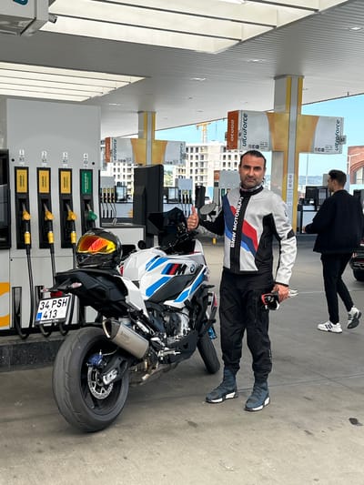 A man wearing a BMW Motorrad jacket and holding a helmet is standing beside a BMW motorcycle at a gas station. The gas station advertises "ultraforce" motorin. The motorcycle's license plate reads "34 PSH 413" with the Turkish flag. Another person is standing in the background. The photo was taken at a gas station in Gaziosmanpaşa, Turkey.