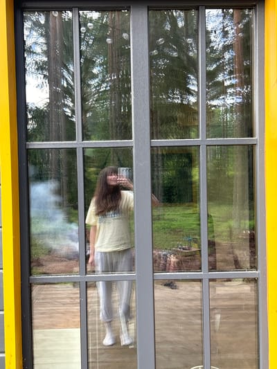 A full-body, mirrored selfie is taken by a person standing indoors and looking out through a large, multi-paned window with yellow framing. The person, with long brown hair and wearing a light yellow t-shirt and gray sweatpants, holds a white smartphone to take the picture. Behind the person, the window reflects a forest scene with tall trees. A faint wispy smoke or fog is visible in the reflection to the left of the person. The foreground shows a wooden deck floor.