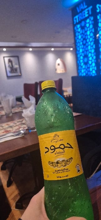 A hand holds a green bottle with a yellow label featuring Arabic text. The bottle appears to contain a beverage. The background includes a wall with decorative lighting and a framed picture. A table with a patterned surface and tissues is visible, along with maroon chairs. A dark screen is mounted higher on the wall. The overall setting suggests an indoor dining area.