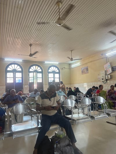 The image shows the interior of a waiting area, possibly a transportation hub, in Mangaluru, India. Several rows of metal chairs are occupied by people of varying ages and attire. Some are using cell phones. There are ceiling fans and arched windows along one wall. The ceiling is paneled. Signs are posted on the wall.