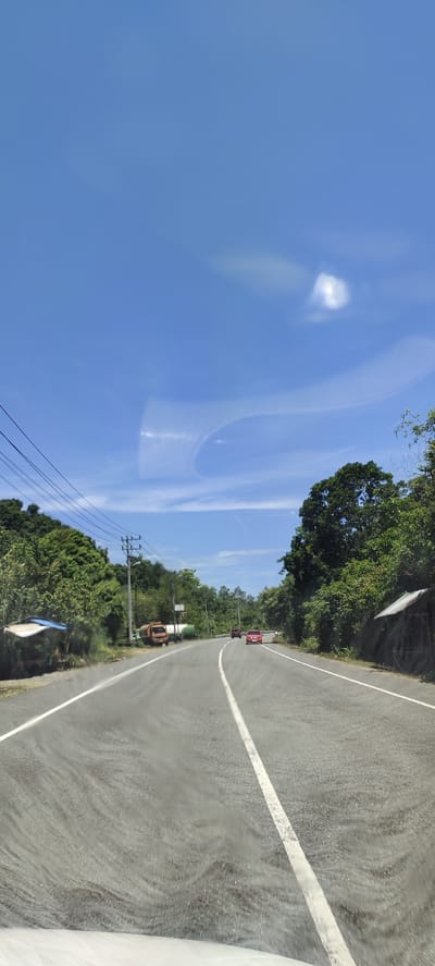 The image depicts a scenic view of a two-lane asphalt road winding through a lush, green landscape. The road is bordered by dense foliage and tall trees, with power lines running along the left side. The sky is a vibrant blue, with a few wispy clouds and a bright, sunlit area, suggesting a clear day.

In the distance, two red cars are visible, traveling in the same direction as the camera. On the left side of the road, there is a small, open-sided structure with a blue roof, and further along, a truck with an orange cab and a white trailer. On the right side of the road, a small, rustic hut with a metal roof is nestled among the trees.

The image appears to have been taken from a moving vehicle, as indicated by the slight blur and the perspective of the road. The overall impression is one of a peaceful, rural setting, likely in a tropical region given the vegetation.

While the prompt mentions the "city of Pulot, Indonesia," the image itself does not contain any specific geographical markers, such as signs or landmarks, that would definitively confirm this location. The scene is consistent with many rural or semi-rural areas in Southeast Asia, but without additional information, the specific location cannot be confirmed from the visual data alone.