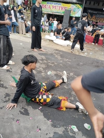 The image depicts an outdoor public gathering on an asphalt road in Sumberejo, Indonesia.

In the immediate foreground, a young male is seated on wet, dark asphalt, leaning back on his left hand. He wears a black long-sleeved shirt with a white logo, dark pants featuring a prominent multi-colored checkered pattern (black, yellow, red, white) around the thighs and knees, and a yellow-red fringed trim at the shins. A green fabric object is visible beneath his lower back. He wears white and grey sneakers with black laces. Scattered dark purple petals and several small clear plastic bags/objects are visible on the asphalt around him.

To the left, a tall, barefoot young male stands facing right, wearing a black long-sleeved shirt and black wide-leg pants. Partially visible to his left are the lower legs and white sandals of another individual. In the mid-ground, several people are present. One individual in black clothing and sneakers walks away towards the right. Others are seated on white bundles, possibly sandbags, along the roadside, facing generally towards the right.

The background features a commercial building with a large green and yellow banner for "WARUNG SEMBAKO KEDUA PUTRI," displaying an address (RT. 03, RW. 10) and a phone number (0838 3411 888). To the right of the building, a large black speaker unit is visible, along with a partial banner for "ALVIN MUSIK." A crowd of spectators, including adults and children, stands on an elevated embankment to the left and congregates near the building to the right, observing the event. The environment suggests an outdoor festival or performance under diffused daylight conditions.