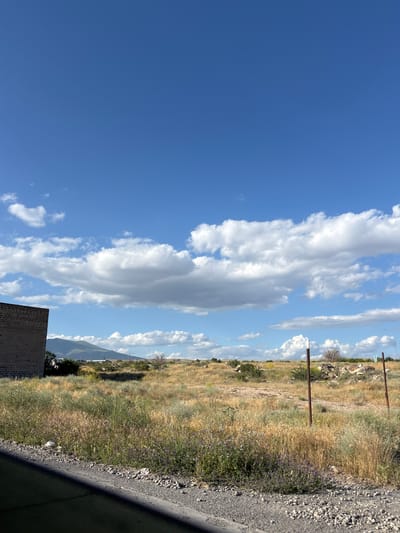 This is a photograph of a rural landscape under a bright, blue sky with scattered white clouds. The scene is dominated by dry, golden-brown grass and vegetation, suggesting a warm, possibly arid climate. In the foreground, to the right, there is a rough gravel surface that appears to be a roadside or pathway.
In the middle ground, a field stretches out, dotted with what appear to be small purple wildflowers mixed in with the dry grass. There are several thin, dark brown posts scattered across the field, which could be remnants of fencing or part of a utility line. In the background, a mountain range is visible, with slopes covered in vegetation. A brick structure, partially visible at the bottom left, adds an element of human construction to the natural setting.
The lighting suggests it is daytime, likely late morning or early afternoon, given the clear sky and bright illumination of the landscape. There are no people visible in the image, nor any obvious activity taking place. The overall impression is one of a peaceful, somewhat desolate natural environment. The specific location context provided is Proshyan, Armenia. There is no visible text in the image.