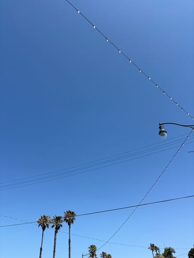 The image shows a low-angle view of a clear, bright blue sky in Redondo Beach, California, United States. Several elements are suspended against the sky: strings of decorative lights with small, round bulbs, a single streetlight with a glass bulb cover, and various power lines. Palm trees are visible along the right edge of the image.