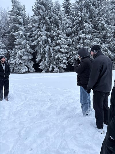 Four males are present in a snow-covered outdoor environment in Le Hohwald, France. In the background, numerous evergreen trees are heavily laden with snow, set against an overcast sky. The ground is entirely covered in thick, uneven snow.
On the far left, a male subject stands, facing right and slightly smiling, wearing a dark winter jacket, a black beanie, dark pants, and gloves. His shoes have red accents.
To his right, a second male subject stands, facing left and partially away from the camera. He wears a dark hooded winter jacket, light blue jeans, and dark gloves. He holds a white rectangular electronic device, consistent with a smartphone, horizontally with both hands, positioned as if capturing an image or video.
Adjacent to him on the right, a third male subject stands, also facing left and partially away from the camera. He wears a dark beanie, a dark winter jacket, and dark pants. His face is partially obscured by the second male.
On the far right, only the lower torso and legs of a fourth male subject are visible, clad in dark athletic pants and white athletic shoes with dark accents. This subject is also facing left.
