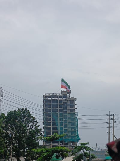In Aminbazar, Bangladesh, a partially constructed building stands against a cloudy sky, its skeletal frame evident. The building is draped in green netting, suggesting ongoing construction. At the very top, a flag with horizontal bands of green, white, and red, and an emblem in the center, flies prominently. Electrical wires crisscross the scene, supported by utility poles, indicating an urban or semi-urban environment. Lush green trees are visible to the left and in the foreground, adding a natural element to the cityscape. The overall impression is one of development and ongoing urban expansion.