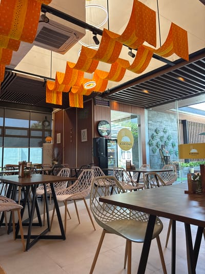 The image depicts the interior of a cafe named KOKONO Cafe & Eatery in Gajahan, Indonesia. The cafe features a modern design with an open layout. The ceiling is adorned with decorative orange and yellow fabric hangings, interspersed with circular light fixtures and an air conditioning unit. Dark wooden beams are visible on the ceiling.
The cafe is furnished with several tables, mostly square and rectangular, with dark wooden tops and black metal frames. There are numerous white plastic chairs with a unique geometric design. Some tables are set with small items like salt and pepper shakers, and a wooden stand holding what appears to be a menu or bill.
Large glass windows and doors offer views of the exterior, suggesting a bright and airy atmosphere. Through the glass, an outdoor seating area with greenery can be partially seen. Inside, a large black speaker and a clock with Roman numerals are mounted on a wall, which is paneled with dark wood. Adjacent to the speaker is a glass-doored refrigerator unit. A prominent circular sign with the KOKONO logo, depicting a steaming cup, is visible on the glass entrance and also on a yellow sign outside. The overall impression is a clean, contemporary, and welcoming dining space. There are no people visible in the image.