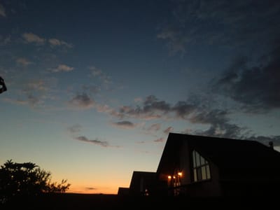 This media file shows a sunset scene with several A-frame style buildings silhouetted against the darkening sky. The sky transitions from a soft orange near the horizon to a deep blue overhead, with scattered clouds visible. The buildings appear to be residential or perhaps a small complex, with some windows faintly illuminated from within. The architecture is distinctive, with steep roofs and triangular window sections. In the foreground on the left, the silhouette of a tree with dark foliage is visible. The overall mood is peaceful and serene, suggesting the end of a day in a quiet, possibly rural, location. The time of day is clearly sunset. There is no visible text.