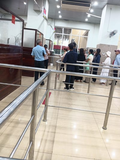 In a brightly lit indoor setting, likely a public service office or government building in Ben Cau, Vietnam, a queue of individuals stands behind metal stanchions. The floor is tiled in a beige, square pattern, and the walls are painted a light color.

In the foreground, a man in a light blue shirt and dark pants stands facing away from the camera, talking to someone out of frame. To his right, a person wearing a light pink shirt and dark pants looks at their phone. Further back in the queue, a woman in a dark top and a light green scarf faces forward, while others in the line are also facing the front.

To the left of the image, a dark wooden counter with glass partitions suggests a service desk area. A red sign with white text is visible on one of the glass panels. In the background, large windows reveal a glimpse of the outdoors. A ceiling-mounted air conditioning unit is visible overhead. The overall impression is one of an orderly queue awaiting service.
