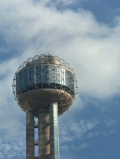 The image features the upper section of a distinctive spherical architectural structure set against a bright, partially cloudy blue sky. The sphere is encased in a geometric, geodesic-style metal framework, within which numerous glass panels reflect the sky and light. Portions of the sphere beneath the metal lattice appear to have a solid, possibly metallic or gold-toned surface.
Extending from the right side of the sphere, a horizontal, glass-enclosed walkway connects it to a broad, concrete structure. This concrete element, which appears to be part of a larger tower, exhibits robust construction with visible supporting sections. The photograph is taken from a low angle, looking upwards, emphasizing the grandeur and elevation of the building.
The scene is set on a clear day, characterized by a vibrant blue sky dotted with scattered white clouds. No people or visible text are present in the frame. Given the location context of Dallas, United States, the spherical structure is identifiable as the observation deck of the iconic Reunion Tower. The image highlights the modern design and geometric complexity of this landmark.