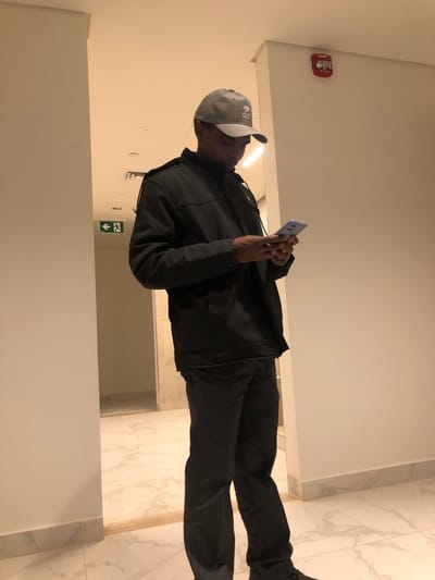 A man in a dark jacket and pants stands indoors, glancing at a blue smartphone. He wears a gray baseball cap. An emergency exit sign with a green arrow and a red fire alarm are visible on the off-white walls. The floor is white marble. The man appears to be in a hallway.