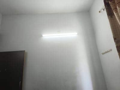 The image shows the interior of a room in Dhandhuka, India. The walls are white. A linear fluorescent light fixture is mounted horizontally near the top of the frame. A dark-colored wardrobe is partially visible in the lower left corner. A curtain with a brown and beige pattern hangs on the right side of the frame. A small row of hooks is mounted on the wall near the curtain.