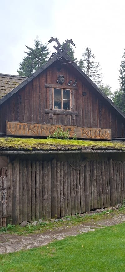 The image shows a wooden building with a moss-covered roof, located in Saula, Estonia. The building features a decorative wooden sign with carved lettering, two carved dragon heads on the roof ridge, and a small window. The facade is constructed from vertical wooden logs. Green grass and a dirt path are visible in the foreground. The sky is overcast.