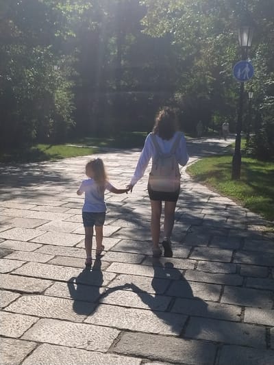 A woman and a young girl are walking away from the camera on a paved path in a park. The woman is wearing a white long-sleeved shirt, a black skirt, and a backpack. The girl is wearing a light blue t-shirt and denim shorts. They are holding hands. Sunlight filters through the trees, casting long shadows on the path. A lamppost with a pedestrian crossing sign is visible to the right of the path. The park appears to be a well-maintained area with grass and trees lining the path. Based on the presence of the pedestrian crossing sign and the general appearance of the park, this scene is likely located in a city park in Písek, Czechia.