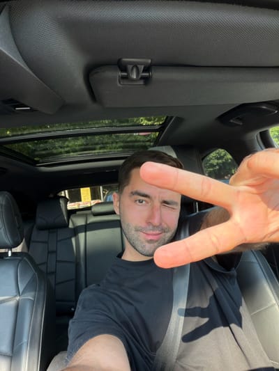 A man is taking a selfie in the driver's seat of a car. He is wearing a black t-shirt and has his seatbelt on. He is winking and holding up a peace sign with his right hand. The car has black leather seats and a sunroof. Through the sunroof, trees and a building can be seen. The scene appears to be in a sunny outdoor environment. There is no visible information in the image that would indicate the location is Cassina Rizzardi, Italy.