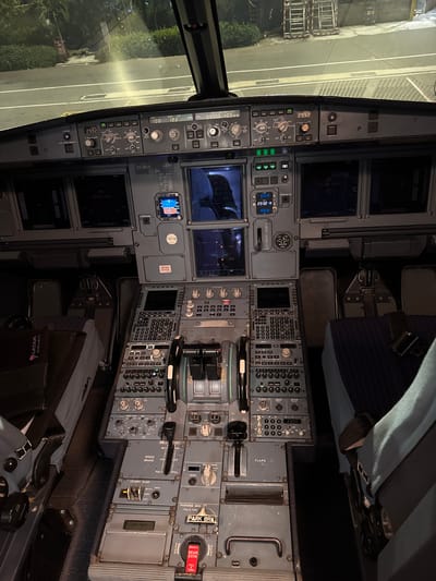 This is a photograph taken inside the cockpit of an Airbus A320 family airliner. The image is taken from behind the seats, focusing on the instrument panel. Various displays show flight information, and the control yoke is visible. The aircraft appears to be on the ground, as seen through the windshield, depicting a runway at night.
"LATAM Airlines" is displayed on a bag situated in front of the panel and the tail number visible on the center instrument panel indicates the aircraft is registered in Chile. There is no verifiable information to confirm the image was taken in Punta Cana.