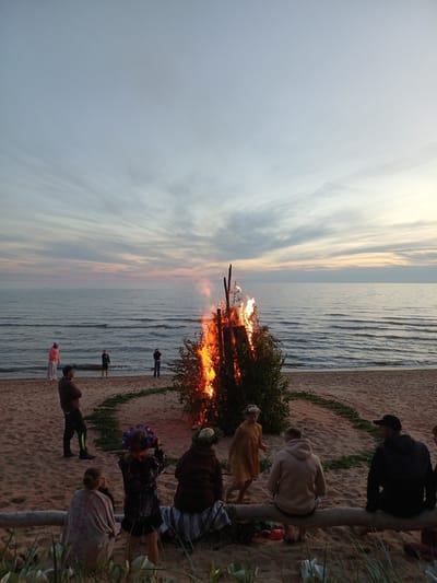 At sunset on a sandy beach near Vārzas, Latvia, a large bonfire blazes in front of the sea. A group of people are gathered on the beach, some standing and watching the bonfire, while others are seated on a log in the foreground. The sky is a gradient of soft pinks and blues, with wispy clouds stretching across the horizon. The sea is calm, with gentle waves lapping at the shore. The bonfire is the central focus of the image, its flames reaching high into the twilight sky. Surrounding the bonfire is a circular arrangement of green foliage, possibly a traditional decoration. The atmosphere appears peaceful and celebratory, suggesting a gathering or festival.