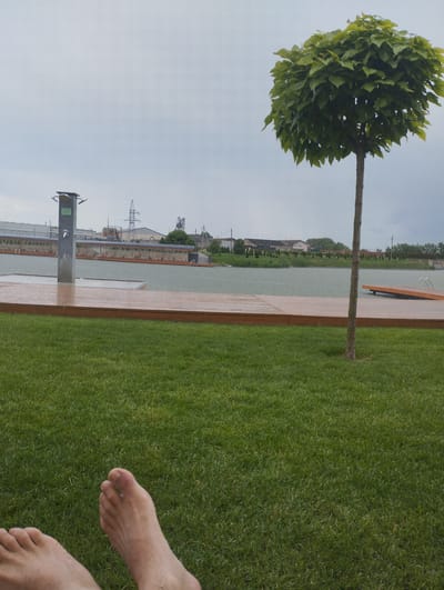 In the city of Romanivka, Ukraine, on a cloudy day, a person's bare feet are visible in the foreground, resting on a lush green lawn. Beyond the lawn, a wooden deck leads to a body of water, which appears to be a swimming area with lounge chairs set up along its edge. A modern outdoor shower fixture stands on the deck. In the background, industrial structures and a tall transmission tower can be seen. A small, pruned tree with a dense canopy of leaves is positioned to the right of the frame, with its slender trunk extending upwards. The water surface shows ripples, suggesting it might be raining or windy.