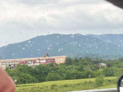 This image captures a panoramic view of the city of Nakaidzeebi, Georgia, under a cloudy sky. The foreground features a lush green field, suggesting a rural or semi-rural setting. In the mid-ground, a large, multi-story building with red and beige facades is visible, likely an apartment complex or a commercial structure, surrounded by dense green trees. Further into the landscape, smaller houses are scattered amongst the trees, hinting at a dispersed settlement pattern. The background is dominated by rolling hills or mountains, covered in vegetation, with a hazy, overcast sky above. Water droplets on the camera lens create a soft, bokeh effect, particularly noticeable in the sky and on the distant hills. The overall impression is one of a tranquil, natural environment with scattered human habitation.